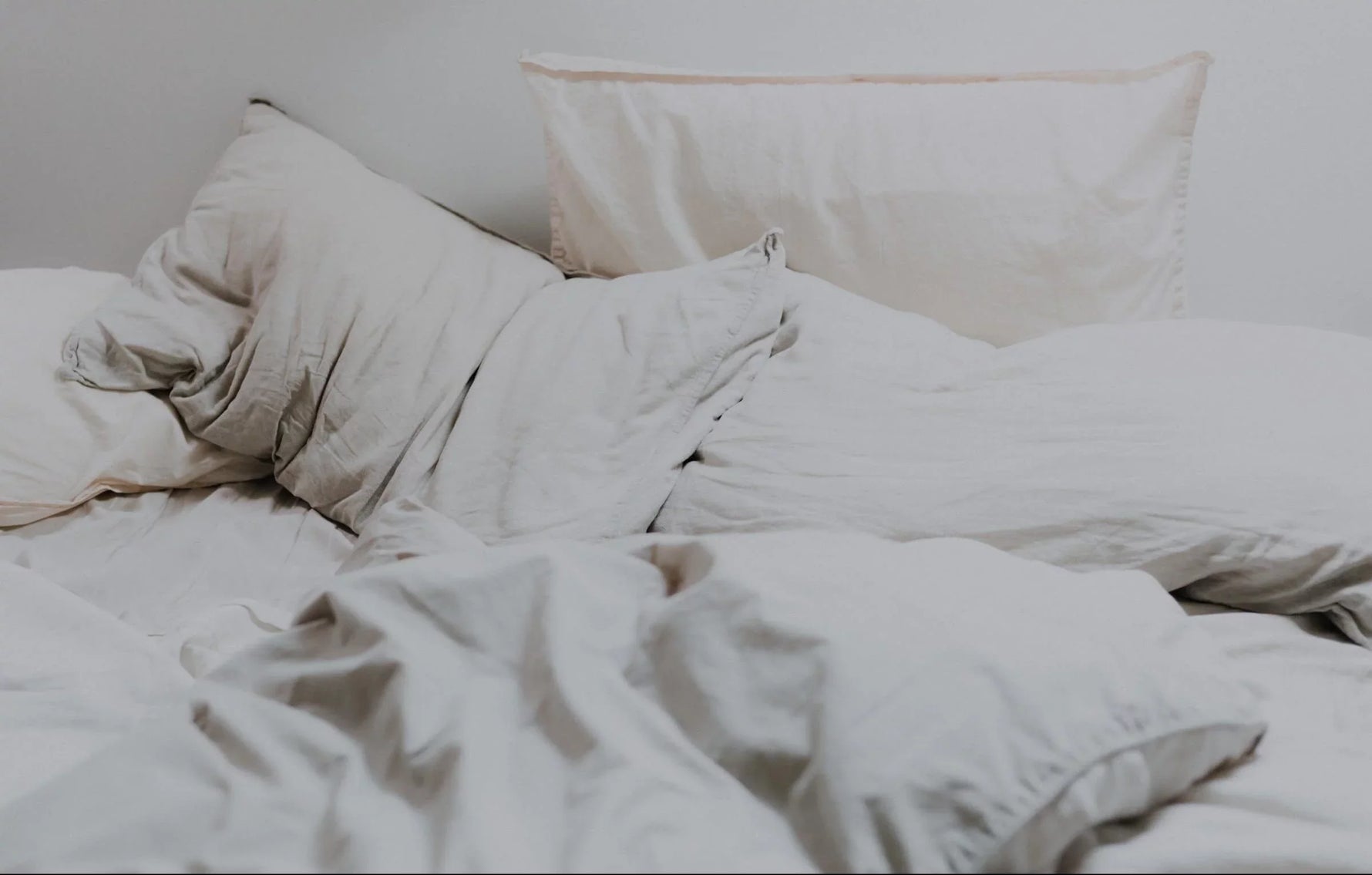 Unmade light beige bedding with rumpled duvet and pillows on a bed against a plain light wall.