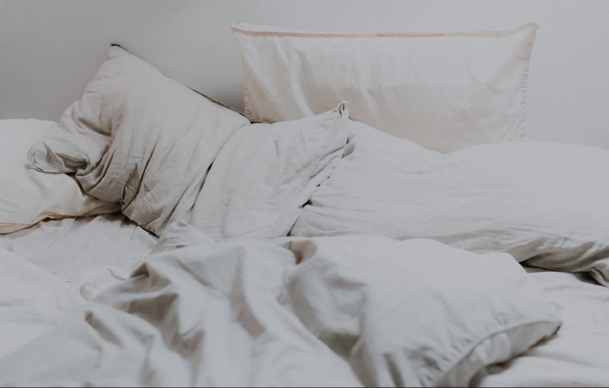 Unmade light beige bedding with rumpled duvet and pillows on a bed against a plain light wall.