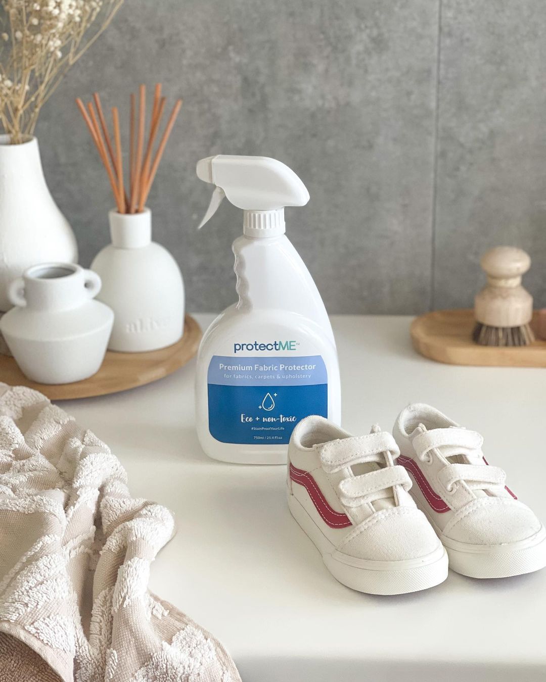 Non-toxic fabric protection spray, protectME, next to a child's or toddler's white sneakers on a surface with decorative items in the background. It demonstrates that protectME can be used to spill proof a kid's shoes and highlights the non-toxic nature of protectME.