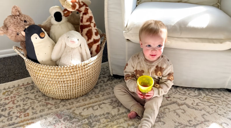 Child holding a yellow cup next to a basket of stuffed animals on a carpeted floor.