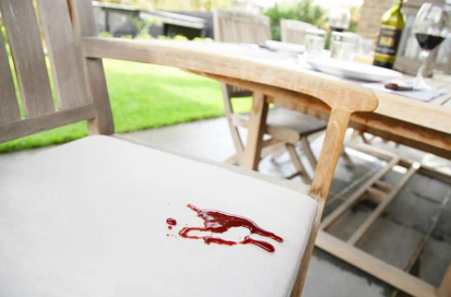 A close-up of a white upholstered chair seat with a red liquid spill or stain, set in a real outdoor patio environment with a wooden dining table and chairs blurred in the background.