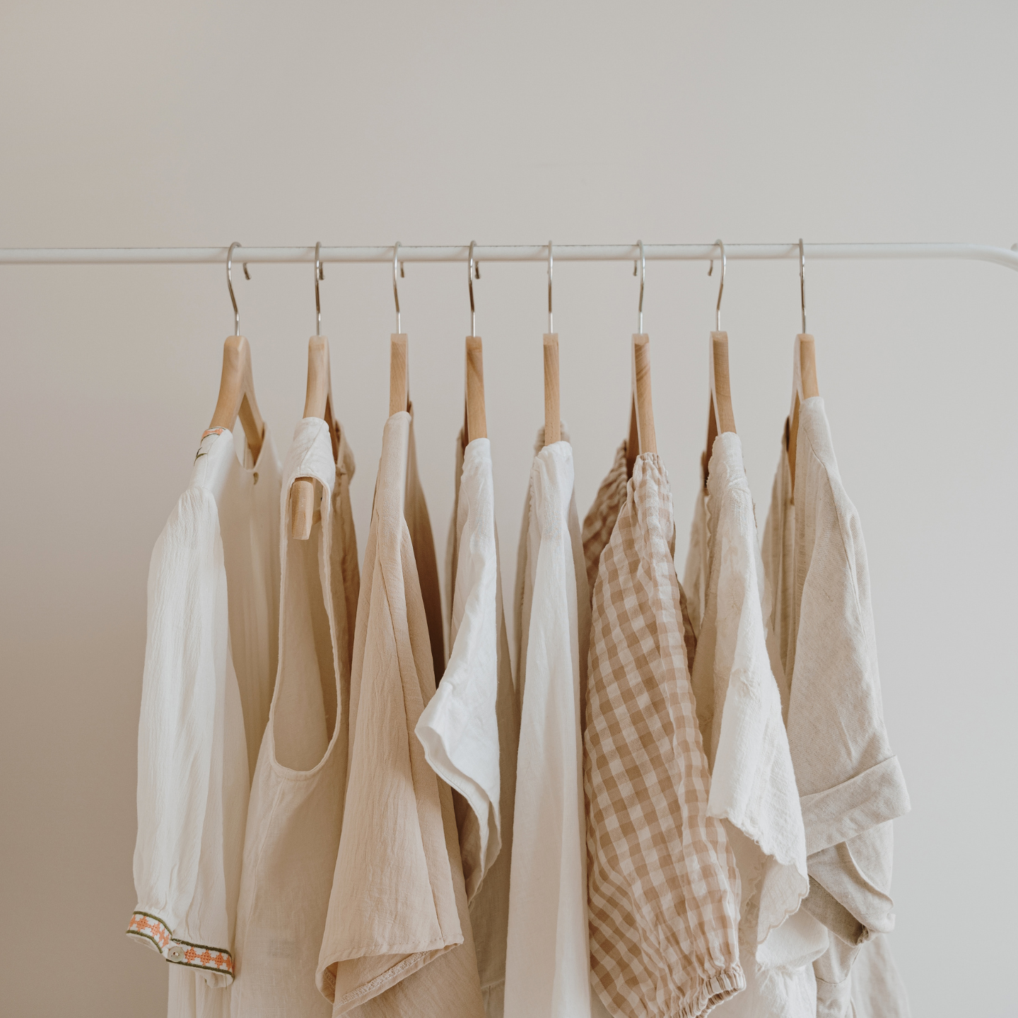 A set of neutral-toned linen and cotton garments hanging on wooden hangers from a thin metal rod against a plain off-white wall.